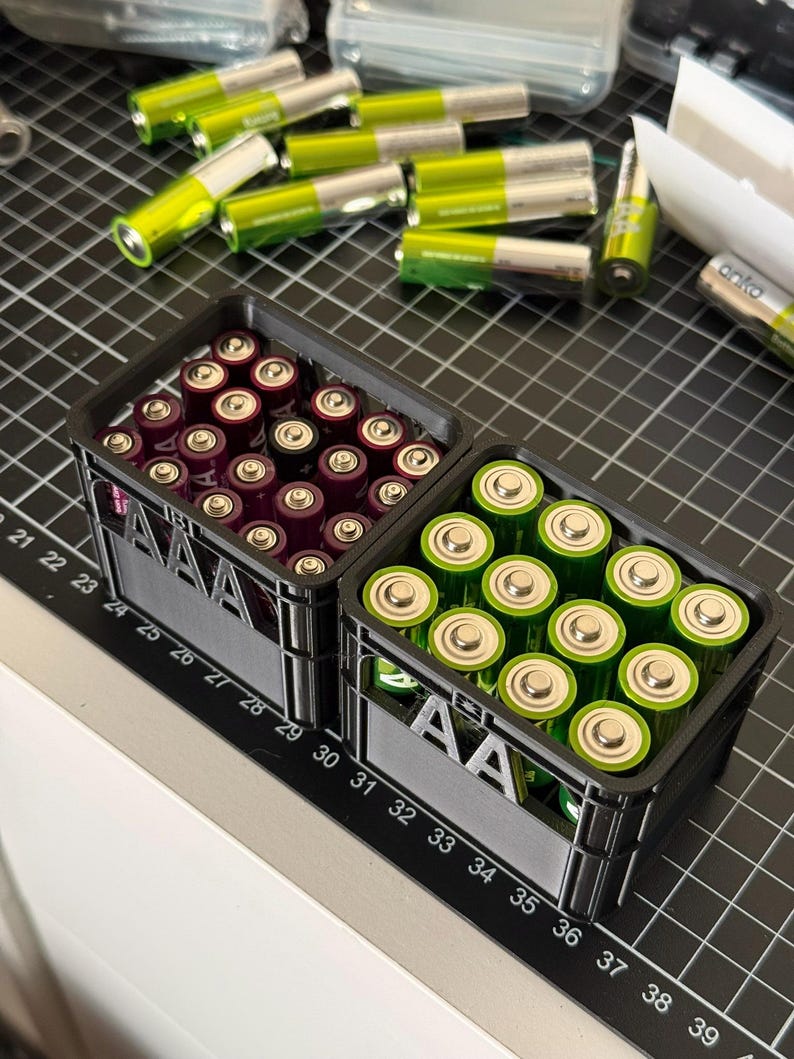 May include: Two black plastic storage containers with the label "AA" on the front. The containers are filled with batteries, one with purple AA batteries and the other with green AA batteries.