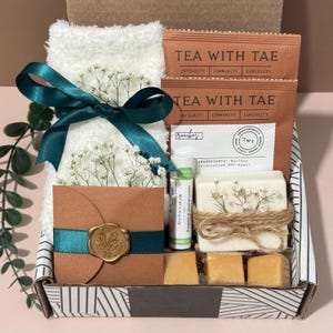 May include: A gift box filled with self-care items. It includes a white towel tied with a teal ribbon, tea packets, a lip balm, soap, and small square soaps. The box has a brown card with a gold seal.