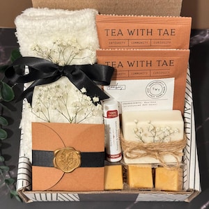 May include: A gift box filled with a white washcloth, tea packets, a lip balm, a bar of soap, and small square soaps. The box is decorated with a black ribbon, dried flowers, and a brown card with a gold seal.