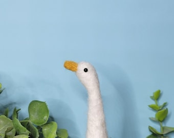 Può includere: Un'oca bianca in feltro ad ago con un becco e piedi gialli. L'oca è in piedi su una superficie di legno con vegetazione verde alle sue spalle.