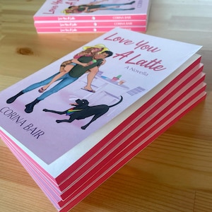 May include: A stack of paperback books titled "Love You A Latte" by Corina Bair. The cover art features a couple and a black dog with a rainbow collar. The book's edges are red, and the background is a light pink.