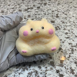 Op de afbeelding: Een lichtgele vilten hamsterfiguur met roze wangen, neus en voeten. De hamster heeft zwarte ogen en zit rechtop. De figuur wordt in een gehandschoende hand gehouden.