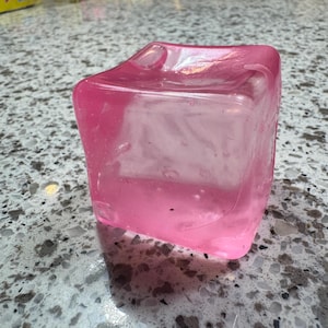 May include: A translucent, pink, square-shaped object with rounded edges. The object has a small, curved indentation on one side. The color is a vibrant, bubblegum pink. The object appears to be made of a solid material.