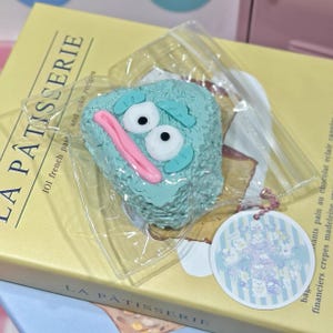 Puede incluir: Un objeto caprichoso en forma de pastel, de color azul claro, con ojos de dibujos animados, una boca rosa y detalles verdes, empaquetado en plástico transparente. El objeto descansa sobre un libro amarillo titulado "LA PÂTISSERIE" con texto sobre pasteles franceses y recetas de pasteles.