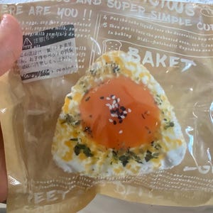 Puede incluir: Una pasta triangular con un centro de color naranja brillante, salpicada con semillas de sésamo negras y blancas, y rodeada por un recubrimiento moteado blanco y verde. La pasta está empaquetada en plástico transparente y papel marrón.