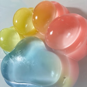 Op de afbeelding: Een close-up van een kleurrijk, hartvormig object. Het object bestaat uit verschillende afgeronde, doorschijnende vormen in tinten geel, oranje, roze en blauw. De kleuren vloeien in elkaar over, waardoor een gradiënteffect ontstaat. Het object lijkt van een gelachtige substantie te zijn gemaakt.