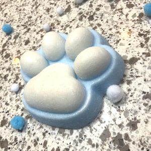 Op de afbeelding: Een lichtblauw en wit pootafdruk-vormig item. De pootafdruk heeft een lichtblauwe basis met witte kussentjes. Kleine witte en blauwe pompons zijn verspreid rond de pootafdruk.