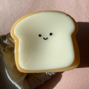 May include: A whimsical, bread-shaped candle with a smiling face. The candle features a white center and a golden-brown crust. The design is simple and cute, perfect for adding a touch of fun to any space.