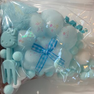 Op de afbeelding: Een doorzichtige plastic zak bevat diverse lichtblauwe en witte decoratieve items. Deze omvatten een Hello Kitty-vormige vork, pompons en wolk-vormige decoraties met glitteraccenten. Een blauw-wit geruite strik voegt detail toe.