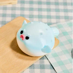 Könnte beinhalten: Hellblaue Kugelfisch-Figur mit schwarzen Augen und einem rosa Mund, auf einem kleinen Holzbrett. Die Figur hat kleine Unebenheiten und Flossen. Der Hintergrund zeigt ein kariertes Muster.