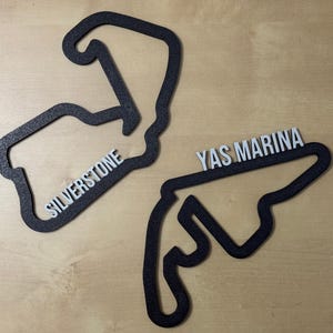 Peut inclure: Deux contours de circuit de course en bois noir, l'un pour Silverstone et l'autre pour Yas Marina. Les contours sont découpés dans du bois et portent le nom du circuit imprimé en blanc.