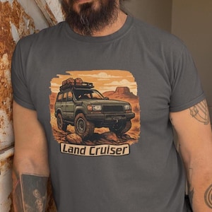 Könnte beinhalten: Dunkelgraues T-Shirt mit einer Grafik eines grünen Land Cruiser SUV in einer Wüstenlandschaft. Das Bild enthält den Text "Land Cruiser". Der SUV hat einen Dachgepäckträger und fährt auf felsigem Gelände. Der Hintergrund zeigt eine Wüstenlandschaft.
