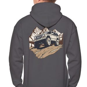 May include: Dark grey hoodie with a graphic of a white SUV driving up a rocky hill, with a mountain range in the background. The design is in shades of brown and tan. The hoodie has a hood and long sleeves.