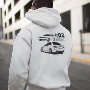 May include: A white hoodie featuring a graphic of two cars and the text "E82 Owner's Club" in black. The hoodie is made of a soft material and has a hood and long sleeves. The image is taken from the back.