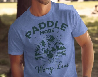 Puede incluir: Camiseta azul con un diseño verde de montaña, kayak y la frase "Paddle More Worry Less". El diseño incluye un sol, pájaros y árboles. La camiseta está hecha de un material suave.