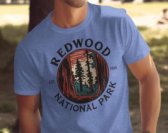 May include: A light blue t-shirt featuring a circular graphic for Redwood National Park. The graphic includes a forest scene with trees and the text "REDWOOD NATIONAL PARK" and "EST. 1968".