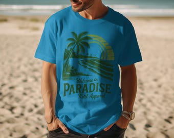 May include: A turquoise t-shirt with a tropical graphic. The design features a palm tree, waves, and a setting sun, with the text "Welcome to Paradise" and "Kittl Apparel" in green. The shirt is worn by a person on a sandy beach.