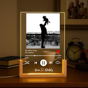 以下が含まれることがあります： 木製のベースが付いた、照明付きのアクリルパネルには、子供を抱いている人の白黒画像が表示されています。 パネルには、エリック・クラプトンの「My Father's Eyes」という曲と「Anna & Daddy」という名前が表示された音楽プレーヤーインターフェースが表示されています。