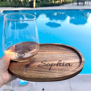 May include: A wine glass with a wooden drink holder. The holder is engraved with the name "Sophia". The glass is filled with a light-colored liquid. The background shows a swimming pool.