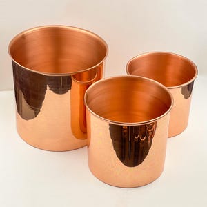 Könnte beinhalten: Drei kupferfarbene Metallbehälter unterschiedlicher Größe. Die Behälter sind zylindrisch mit glatter, reflektierender Oberfläche. Der größte Behälter befindet sich links, mit zwei kleineren Behältern rechts. Die Behälter sind leer und haben eine polierte Oberfläche.