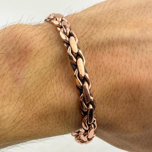 May include: A copper-coloured bracelet with a chain link design. The bracelet is made of interlocking oval links, creating a textured surface. The bracelet is worn on a person's wrist.
