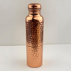 May include: A copper water bottle with a hammered texture. The bottle has a screw-on cap and a cylindrical shape. The copper has a shiny, reflective surface. The bottle is designed to hold liquids and is suitable for everyday use.