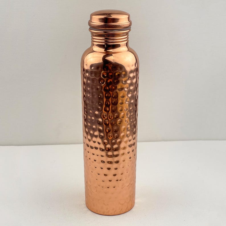 May include: A copper water bottle with a hammered texture. The bottle has a screw-on cap and a cylindrical shape. The copper has a shiny, reflective surface. The bottle is designed to hold liquids and is suitable for everyday use.