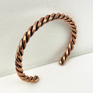 May include: A copper-colored cuff bracelet with a twisted rope design. The open-ended bracelet is made of a shiny, reddish-brown metal. The bracelet is shown against a white background.