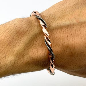 May include: A copper-colored twisted bracelet with silver-colored accents. The bracelet is worn on a person's wrist. The design features a spiral pattern with a textured finish, showcasing a blend of metallic hues.