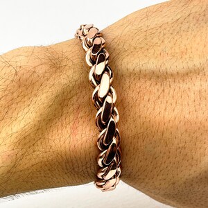 May include: A rose gold chain link bracelet. The bracelet is made of interlocking oval links, creating a thick, textured design. The bracelet is worn on a wrist, showcasing its polished, metallic finish.