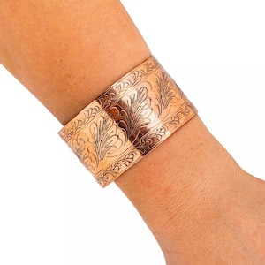 May include: A wide copper cuff bracelet with an engraved floral design. The bracelet is approximately 2 inches wide and features detailed leaf and vine patterns across its surface. The copper has a warm, reddish-brown hue, and the design is etched into the metal.