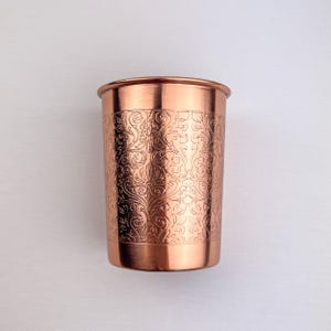 May include: A copper-colored drinking glass with an etched floral pattern. The glass has a cylindrical shape with a slightly flared rim. The etched design covers the majority of the glass's surface, adding a decorative element to the piece.