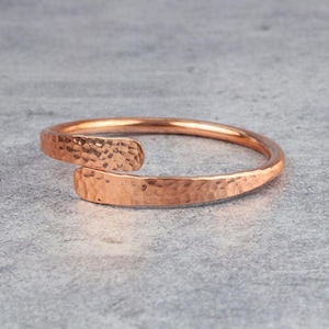 May include: A copper-colored cuff bracelet with a hammered texture. The open-ended design features overlapping ends, creating a modern and minimalist aesthetic. The bracelet is displayed on a gray surface, highlighting its warm metallic tones.