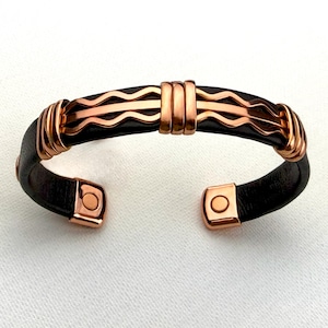 May include: A copper and leather magnetic bracelet with a wavy design. The bracelet has a black leather band with copper accents and magnetic closures.