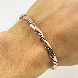 May include: A copper and silver-toned bracelet with a twisted design. The bracelet features alternating sections of copper and silver metal, creating a textured and eye-catching pattern. The bracelet is worn on a wrist.