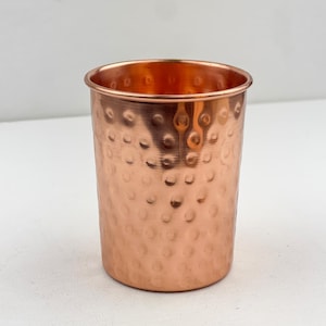 May include: A copper-colored tumbler with a hammered texture. The cylindrical cup has a slightly flared rim and a polished interior. The exterior features a pattern of evenly spaced, small, rounded indentations. The cup is designed to hold liquids.