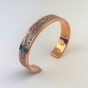 Op de afbeelding: Koperen manchetarmband met een gegraveerd bloemmotief. De armband heeft een groene emaille inleg en drie magnetische sluitingen. Het open ontwerp maakt het gemakkelijk om te dragen. Een stijlvol accessoire.