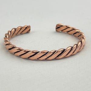 May include: A copper-colored cuff bracelet with a twisted rope design. The open-ended bracelet is made of a solid metal and has a polished finish. The bracelet is approximately 2.5 inches in diameter.