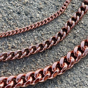 Puede incluir: Tres cadenas de curb de oro rosa de diferentes tamaños. Las cadenas están dispuestas sobre una superficie gris texturizada.