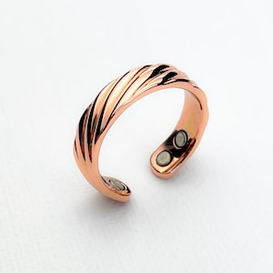 May include: A copper-colored ring with a textured, twisted design. The open-ended ring features two small, round magnets on one end. The ring is shown against a white background.