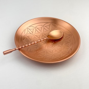 Może przedstawiać: Miedziany łyżka barmańska spoczywa na pasującym okrągłym spodku. Spodek ma wygrawerowany wzór kwiatowy. Łyżka ma skręcony uchwyt i małą, zaokrągloną miseczkę. Przedmioty są prezentowane na białej powierzchni.