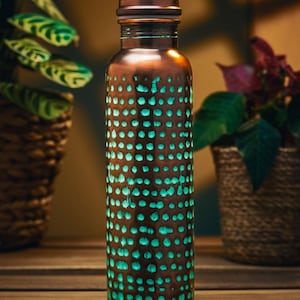 May include: A copper-coloured water bottle with a dotted pattern that glows teal. The bottle is cylindrical with a screw-on cap. The background includes plants in woven baskets, creating a natural aesthetic.