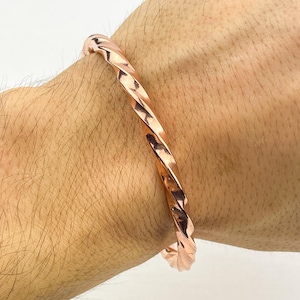 May include: A copper-colored twisted cuff bracelet. The bracelet has a spiral design and is worn on a wrist. The metal has a shiny finish. The bracelet is a piece of jewelry.