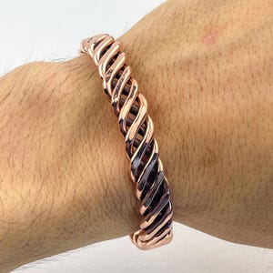 May include: A copper-colored, twisted metal bracelet. The bracelet has a spiral design with alternating light and dark tones. The cuff is worn on a wrist, showcasing the intricate pattern and metallic sheen.
