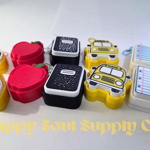 May include: A collection of school-themed silicone keychains featuring red apples, black composition notebooks, yellow school buses, white lined paper sheets, and yellow pencils. The text Happy Soul Supply Co. is printed at the bottom.