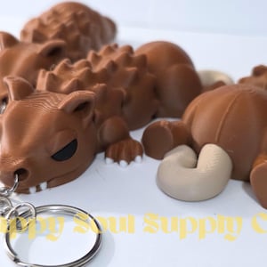 May include: A brown, articulated, 3D-printed ankylosaurus keychain featuring a textured, spiky back, a curled tail, white teeth, and black eyes. The figure is attached to a silver-colored metal split ring. The text Happy Soul Supply Co. is visible.