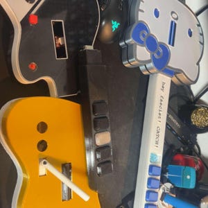 May include: A collection of custom game controllers including a black and white guitar-shaped controller, a yellow guitar-shaped controller, and a white Hello Kitty-themed controller with a blue bow and the text "hey fascist! CATCH!" printed on it.