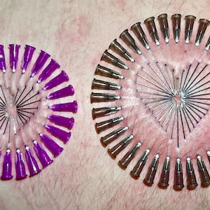 May include: Two circles of needles inserted into skin, forming a heart shape. The needles are arranged in a circular pattern, with the points facing inward. The needles are different colours, one circle is purple and the other is brown.