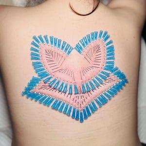 May include: A close-up of a person's back with a flower-shaped arrangement of blue-tipped needles. The needles are inserted into the skin, creating a pink, textured pattern. The arrangement is centered on the back.