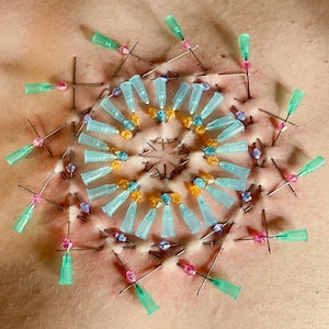 May include: A close-up of a person's torso with a circular pattern of blue, orange, and purple beads and needles inserted into the skin.